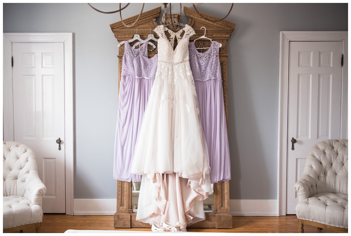 white lace wedding dress hanging with three purple bridesmaids dresses on an ornate mirror at Stone House of St Charles, wedding venue in St Louis Missouri