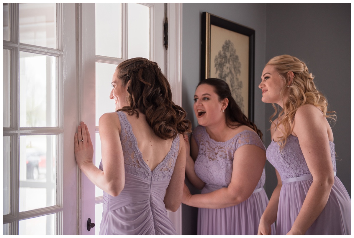 Bridesmaids in lace lilac purple dresses peeking in on bride and groom's emotional first look at Stone House of St Charles, a St Louis wedding venue
