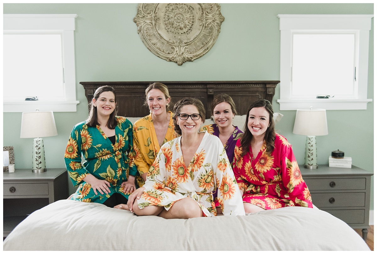 Five women sitting on a bed in multicolor floral kimono-style robes, bride in a white robe with sunflowers