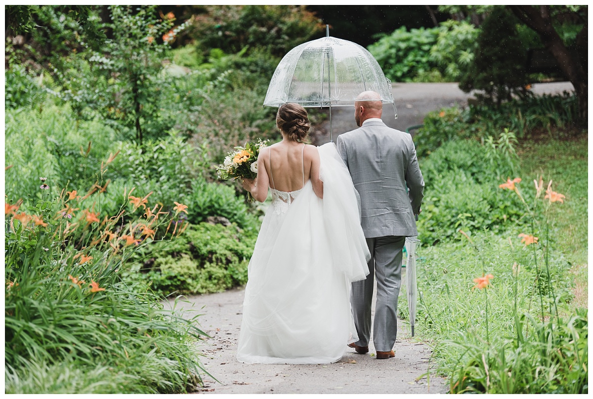 Bride in a tulle-skirted gown and groom in grey suit under an umbrella, walking through the gardens at Lafayette Square Park.