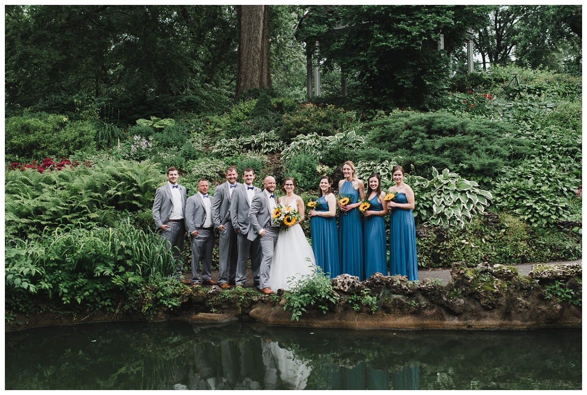 wedding party in grey suits and long blue dresses surrounding bride and groom amid lush green foliage in Lafayette Square Park.