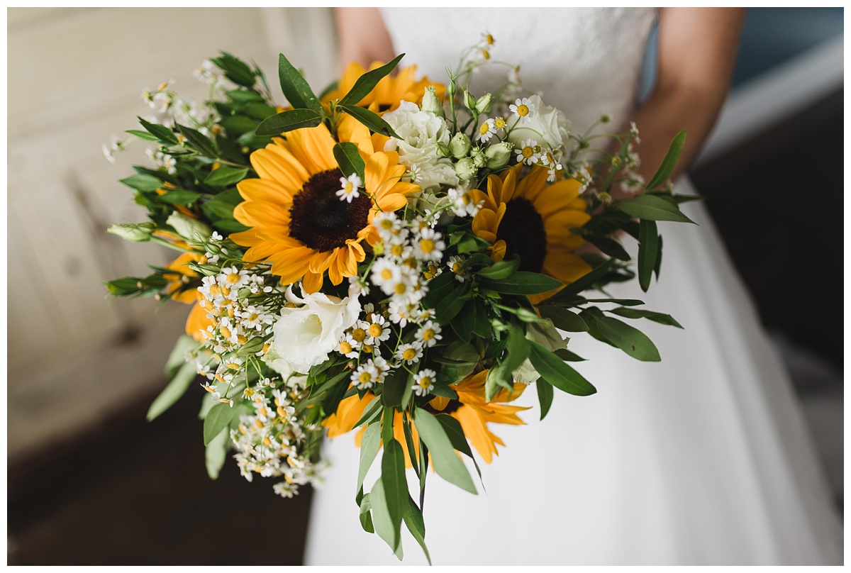 A close-up image of a sunflower bouquet accented with white roses, tiny daisies and greenery